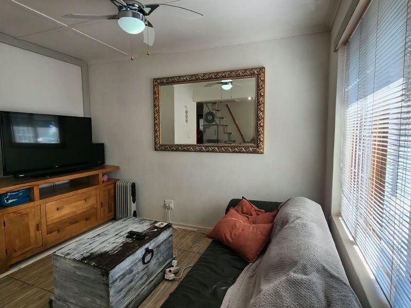 A comfortable living room featuring tiled flooring, a ceiling fan, and large windows fitted with venetian blinds.