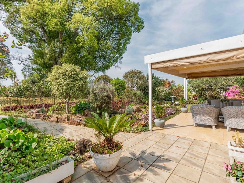 A well-maintained covered patio area featuring tiled flooring, overlooking a lush, landscaped garden with mature trees and stone garden beds.
