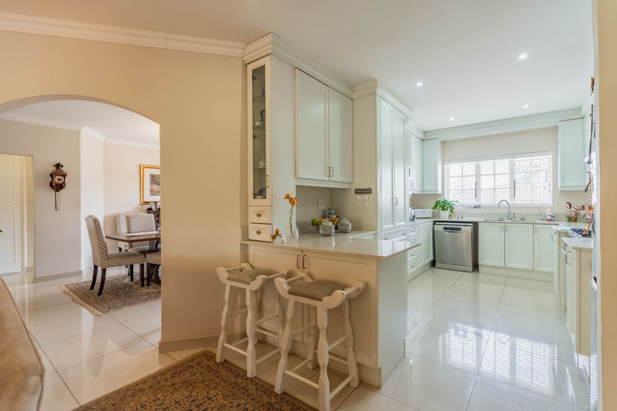 A bright, well-maintained kitchen featuring white cabinetry, stone countertops, a breakfast bar, and tiled flooring, with an open-plan flow to the adjacent dining area.