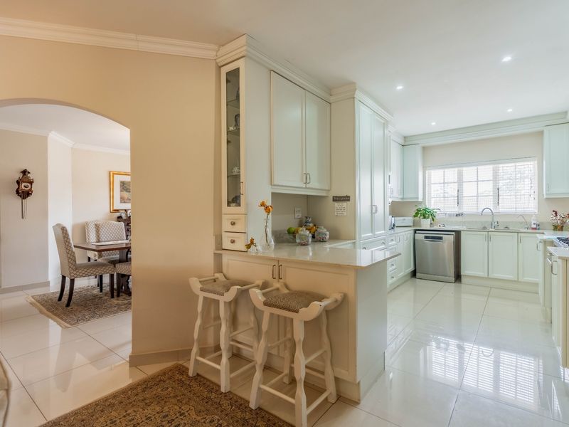A bright, well-maintained kitchen featuring white cabinetry, stone countertops, a breakfast bar, and tiled flooring, with an open-plan flow to the adjacent dining area.