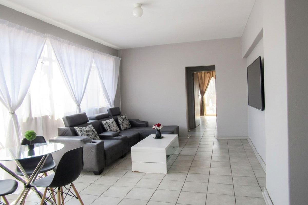 A bright and spacious living room featuring neutral tiled flooring, large windows allowing natural light, and a wall-mounted television.