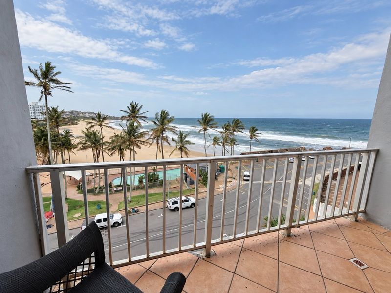 A balcony featuring tiled flooring and a metal balustrade, offering an expansive view of the beach, ocean, and surrounding coastal area.
