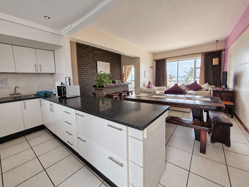 Open-plan living area featuring a modern kitchen with granite countertops, tiled flooring, and a brick feature wall, leading to a bright balcony with views.
