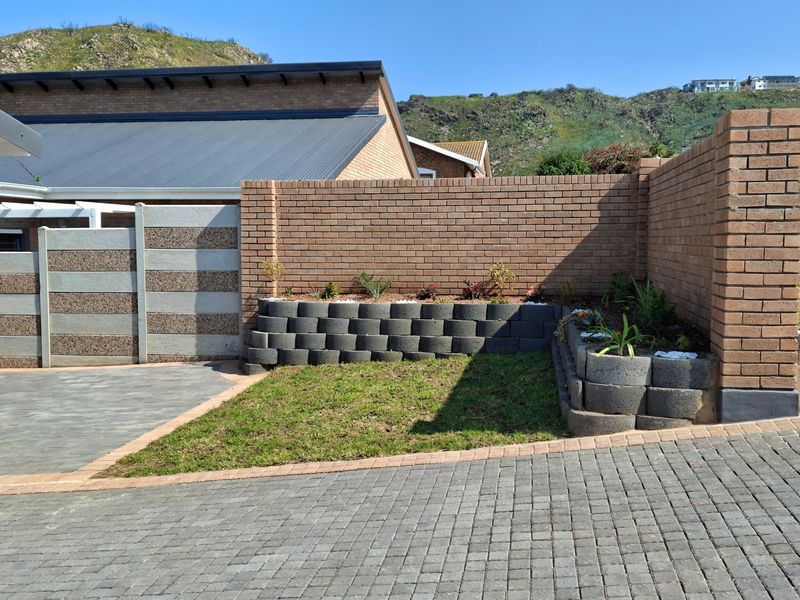A neat front exterior area featuring a paved driveway, brick boundary wall, and tiered garden beds with retaining wall blocks.
