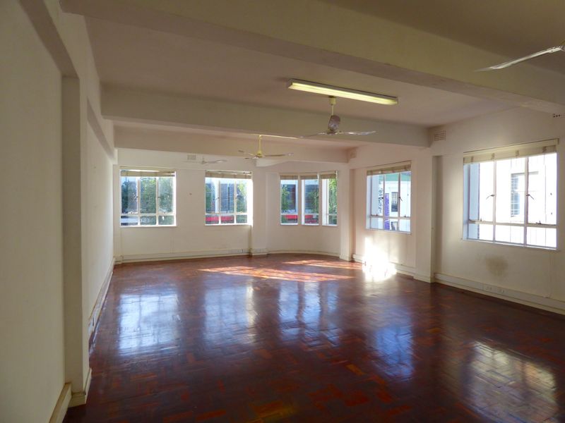 A spacious, empty commercial office area featuring polished parquet flooring, multiple large windows providing natural light, and ceiling-mounted lighting and fans.