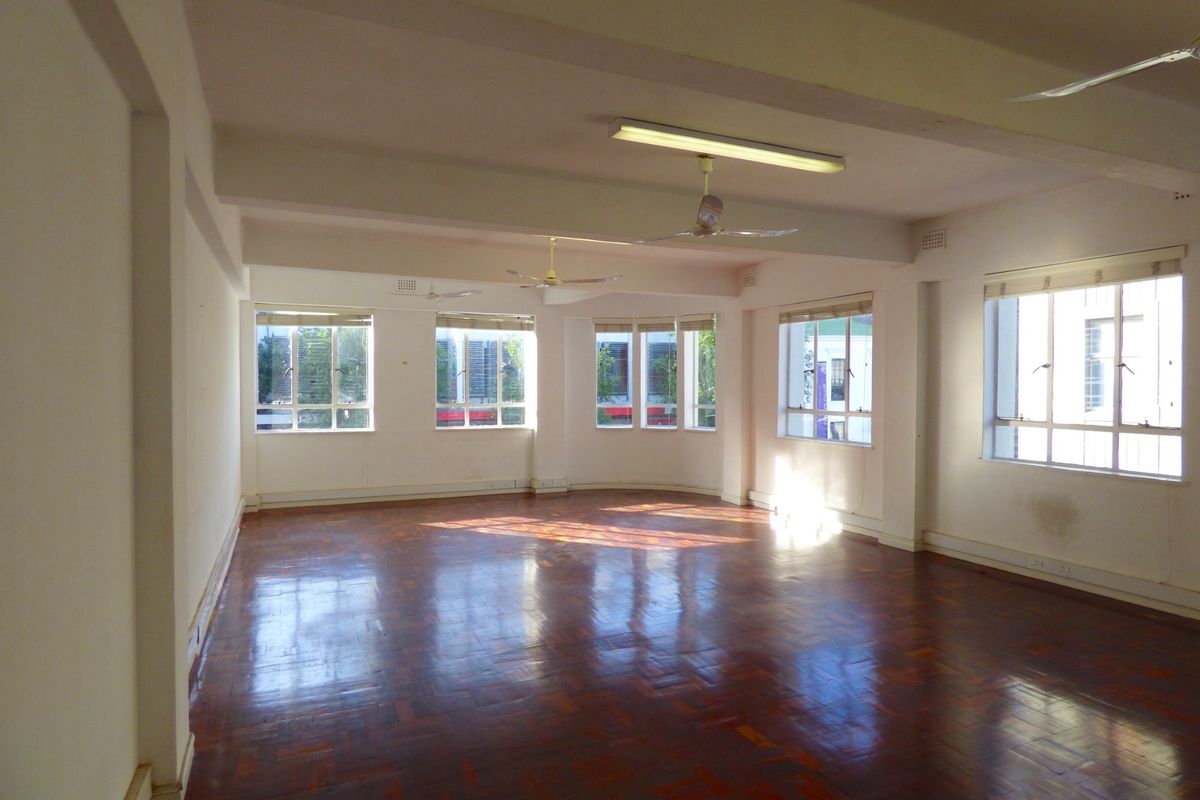 A spacious, empty commercial office area featuring polished parquet flooring, multiple large windows providing natural light, and ceiling-mounted lighting and fans.
