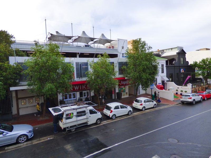 Street-level view of a commercial building featuring retail storefronts, metal security shutters, and a tensile fabric roof structure over the upper level.