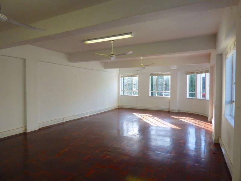 A spacious, empty commercial office area featuring polished parquet flooring, multiple windows providing natural light, and fixed ceiling fans.