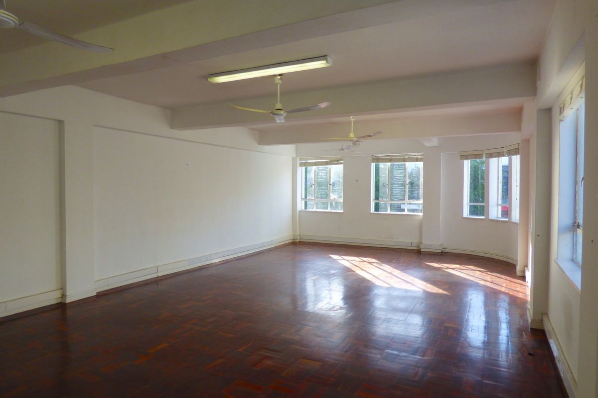A spacious, empty commercial office area featuring polished parquet flooring, multiple windows providing natural light, and fixed ceiling fans.