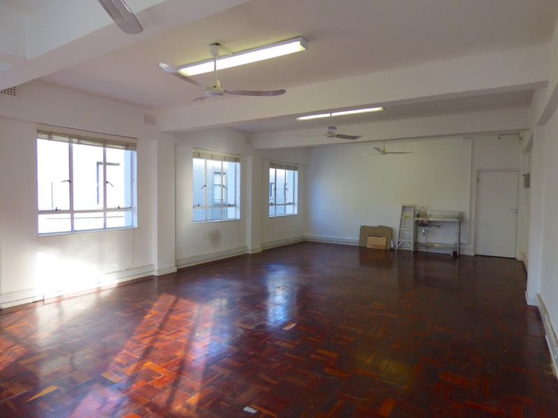 Spacious open-plan commercial area featuring parquet flooring, multiple windows for natural light, ceiling fans, and a basic stainless steel sink unit.
