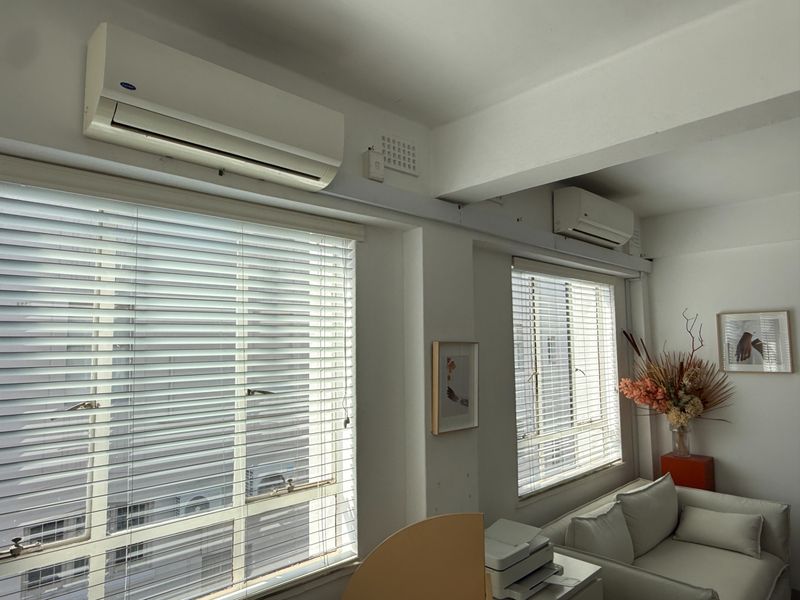 A bright office space featuring dual split-system air conditioning units, large windows with venetian blinds, and an exposed structural ceiling beam.