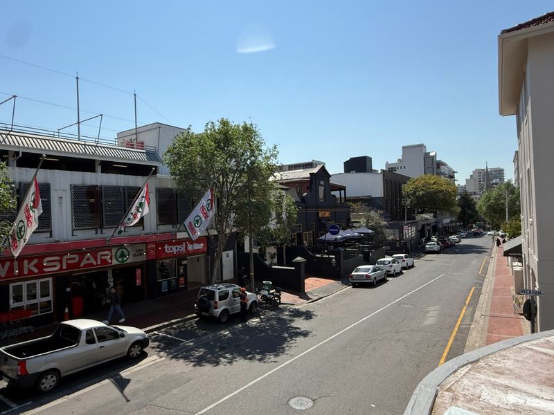 An elevated street-level view of a commercial urban area featuring retail storefronts, paved sidewalks, and street-side parking.