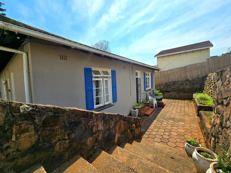 Rear exterior view featuring a paved patio area, stone retaining walls, and a house facade with blue shutters and casement windows.