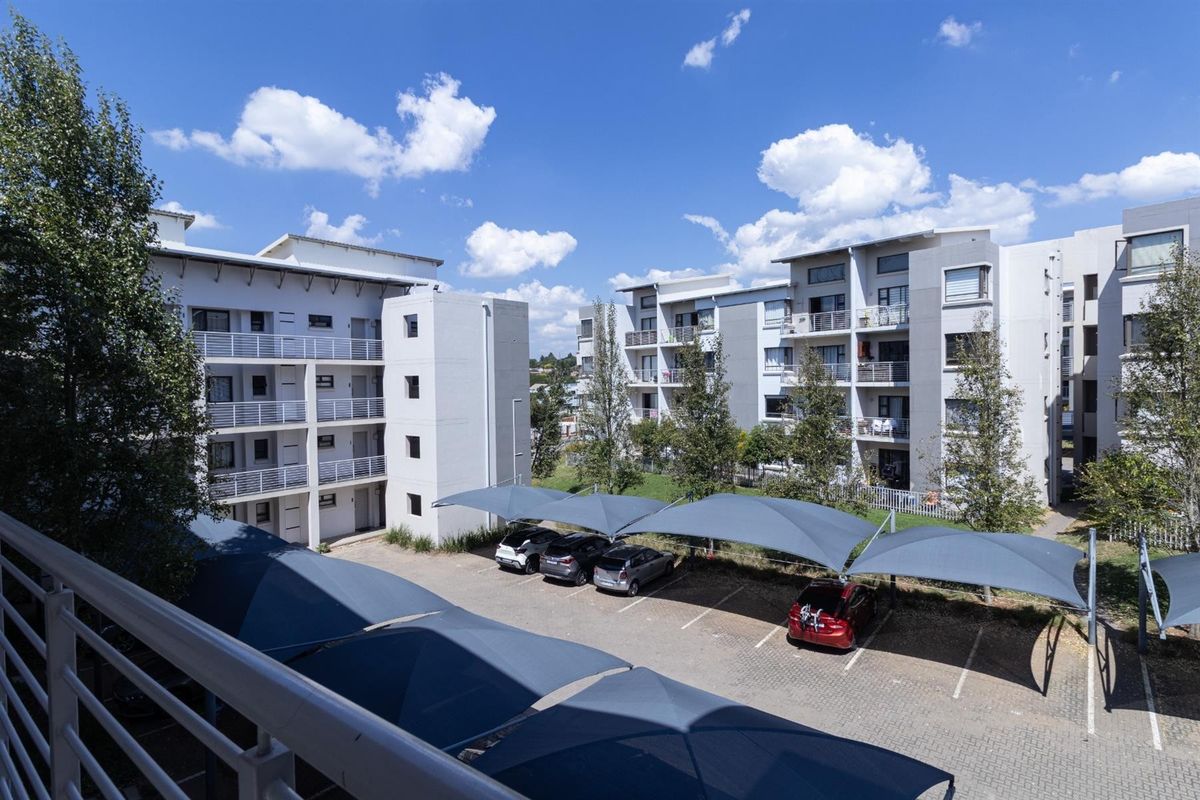 Elevated view of the apartment complex exterior, featuring multi-story residential blocks and a paved parking area with shade sail structures.