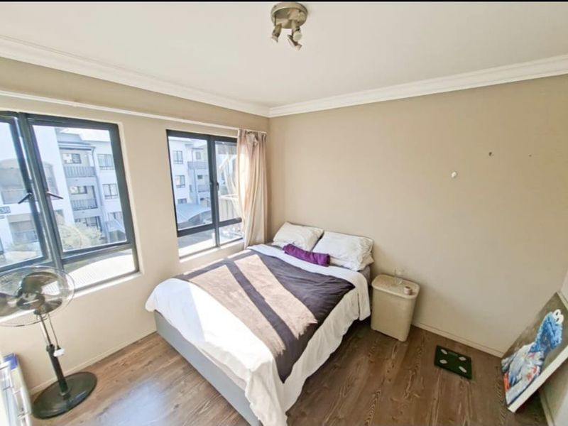 A bedroom featuring laminate flooring and large aluminium-framed windows, providing natural light. The space is functional but would benefit from minor cosmetic wall repairs.