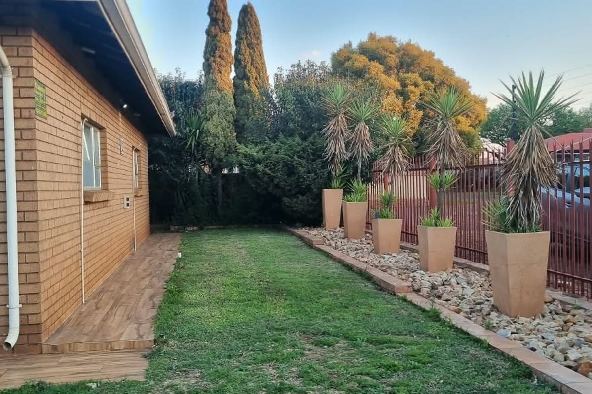 A well-maintained rear exterior view of the townhouse featuring a brick facade, tiled walkway, and a landscaped garden with perimeter security fencing.