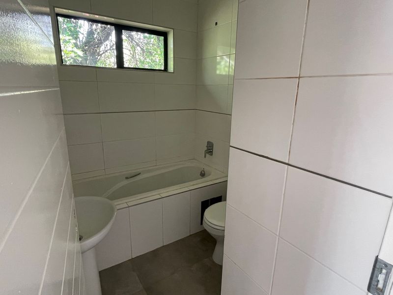 A compact bathroom featuring a bathtub, pedestal basin, and toilet with white wall tiling and neutral floor tiles.