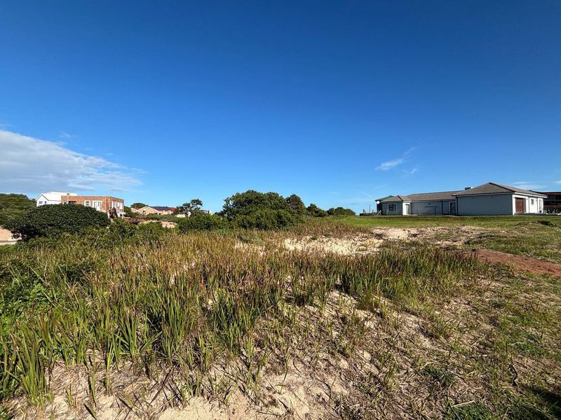 A view of a vacant residential plot featuring natural coastal vegetation and sandy terrain, with neighboring properties visible in the background.
