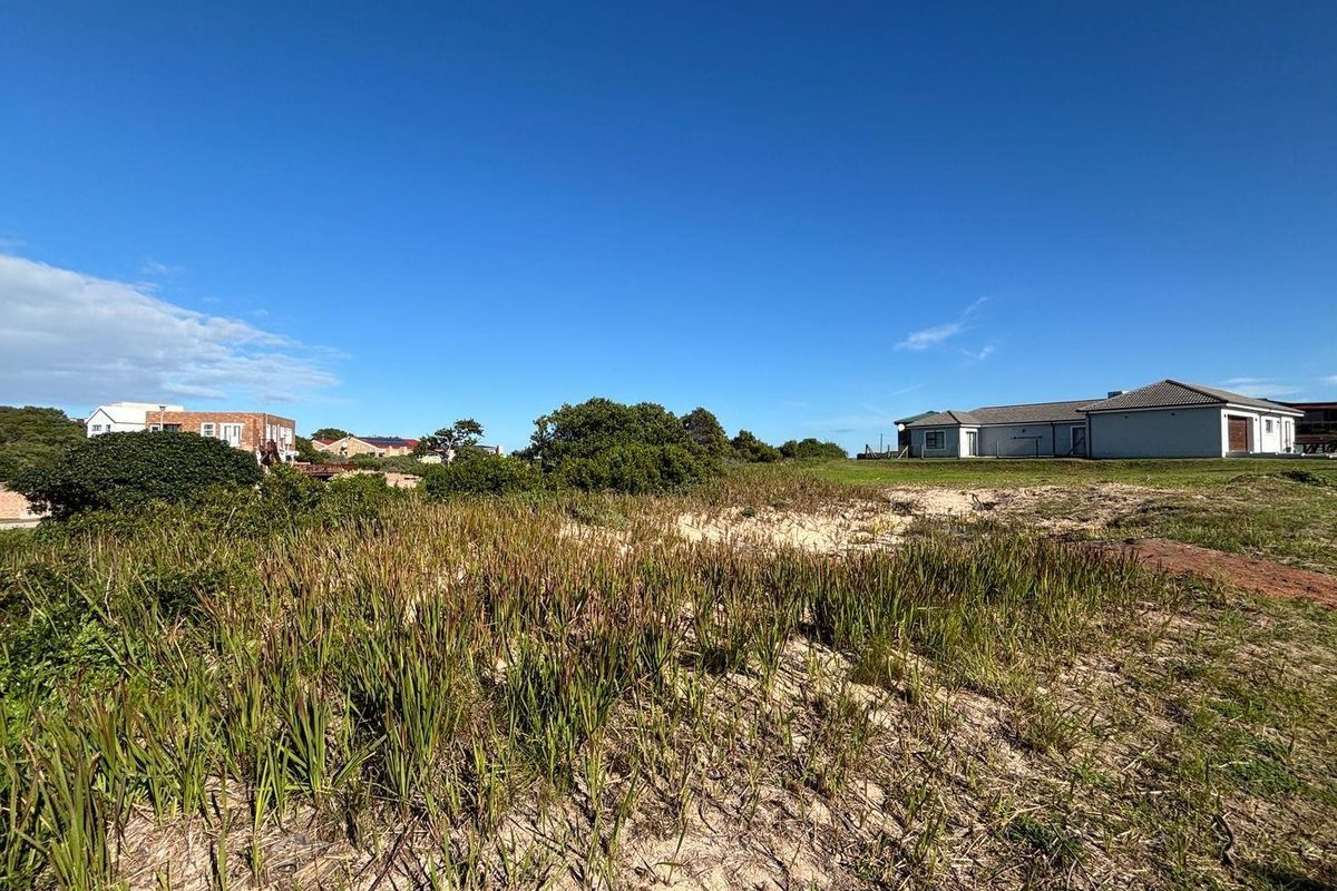 A view of a vacant residential plot featuring natural coastal vegetation and sandy terrain, with neighboring properties visible in the background.
