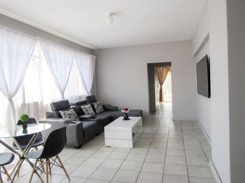 A bright and spacious living room featuring neutral tiled flooring, large windows allowing natural light, and a wall-mounted television.