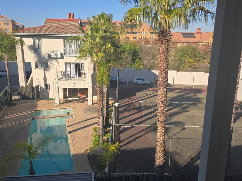 Elevated view of a residential complex featuring a swimming pool, tennis court, and paved outdoor areas surrounded by perimeter walls and palm trees.