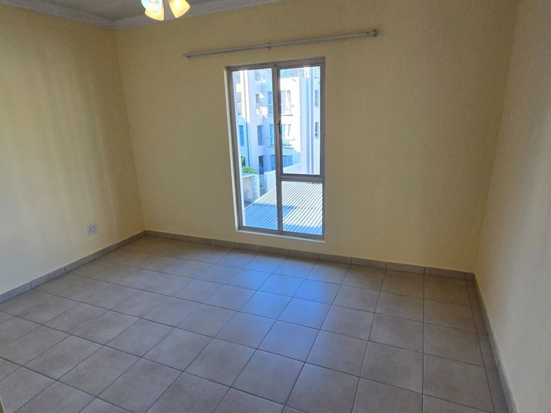 A bright, empty bedroom featuring neutral tiled flooring, a ceiling fan, and a large window with a curtain rod.