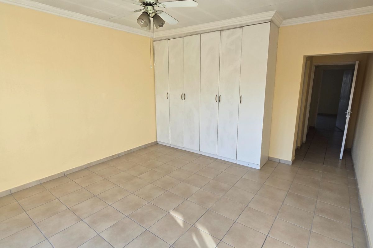 A spacious, empty bedroom featuring neutral tiled flooring, built-in cupboards for storage, and a ceiling fan.