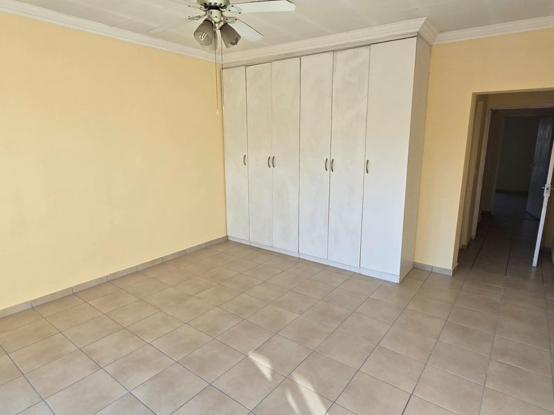 A spacious, empty bedroom featuring neutral tiled flooring, built-in cupboards for storage, and a ceiling fan.