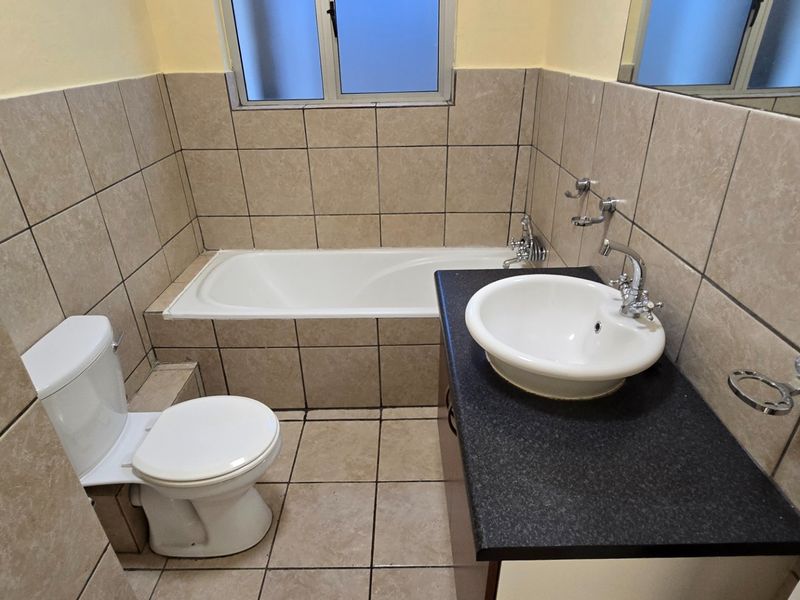 A functional bathroom featuring a built-in bathtub, a vanity basin with a dark countertop, and neutral wall and floor tiling.