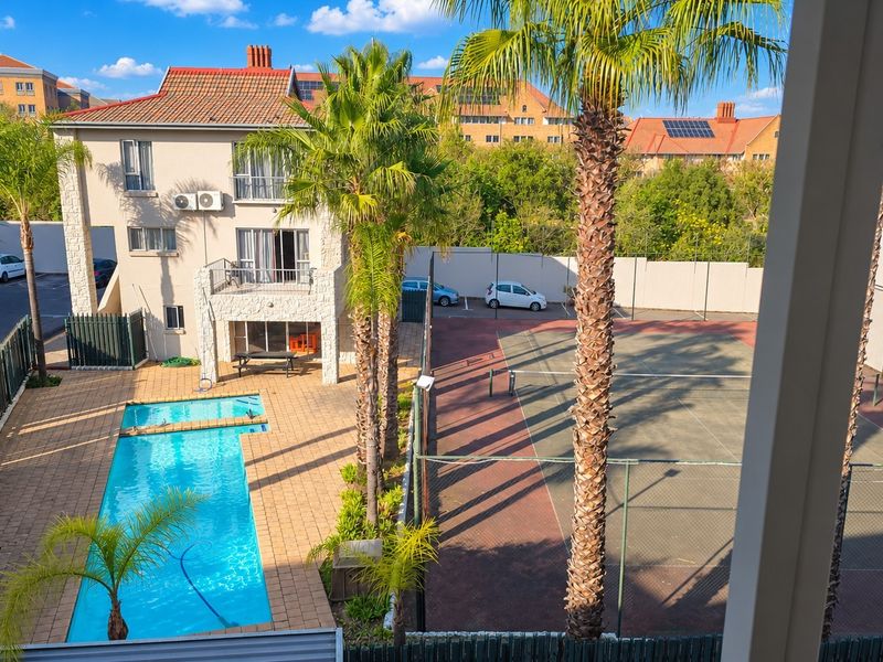 Elevated view of a residential complex featuring a swimming pool, tennis court, and paved outdoor entertainment area surrounded by landscaped palm trees.