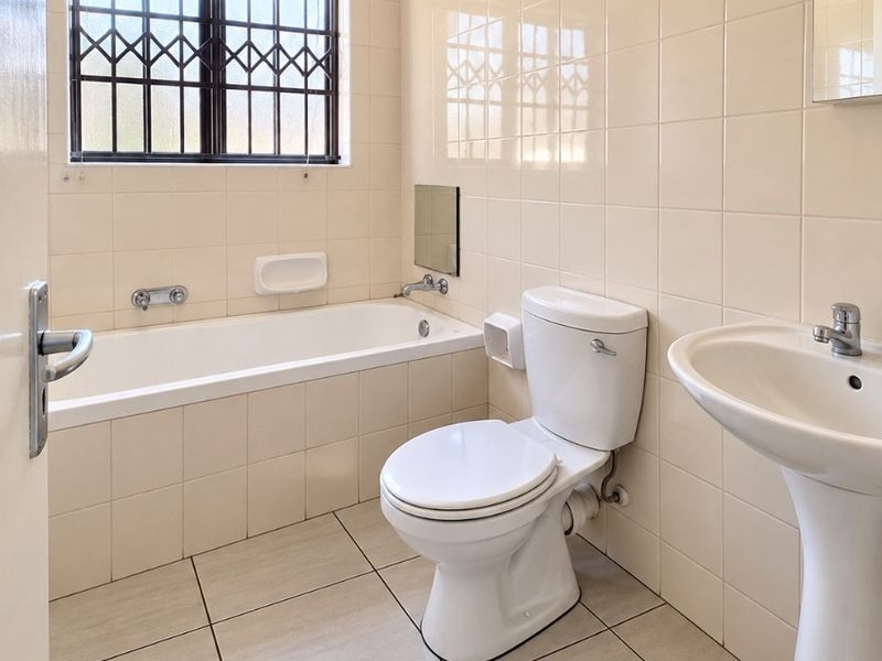 A clean, functional bathroom featuring a bathtub, pedestal basin, and toilet, finished with neutral wall and floor tiles and security bars on the window.