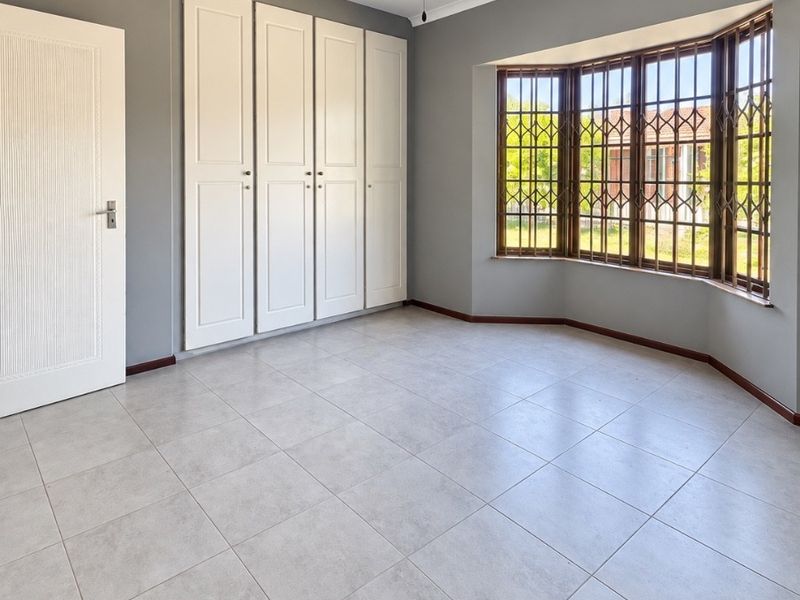 Spacious bedroom featuring neutral tiled flooring, built-in cupboards, a ceiling fan, and a large bay window with security bars.