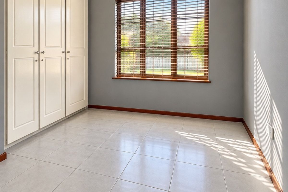 A bright, empty bedroom featuring neutral grey walls, tiled flooring, and a built-in wardrobe with timber venetian blinds on the window.