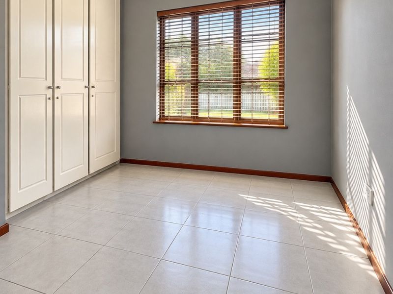 A bright, empty bedroom featuring neutral grey walls, tiled flooring, and a built-in wardrobe with timber venetian blinds on the window.