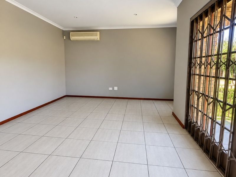 A spacious, empty living area featuring neutral tiled flooring, recessed lighting, and a wall-mounted air conditioning unit with access to an outdoor area via security gate.