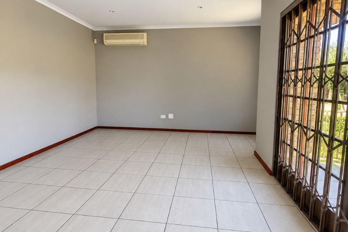 A spacious, empty living area featuring neutral tiled flooring, recessed lighting, and a wall-mounted air conditioning unit with access to an outdoor area via security gate.