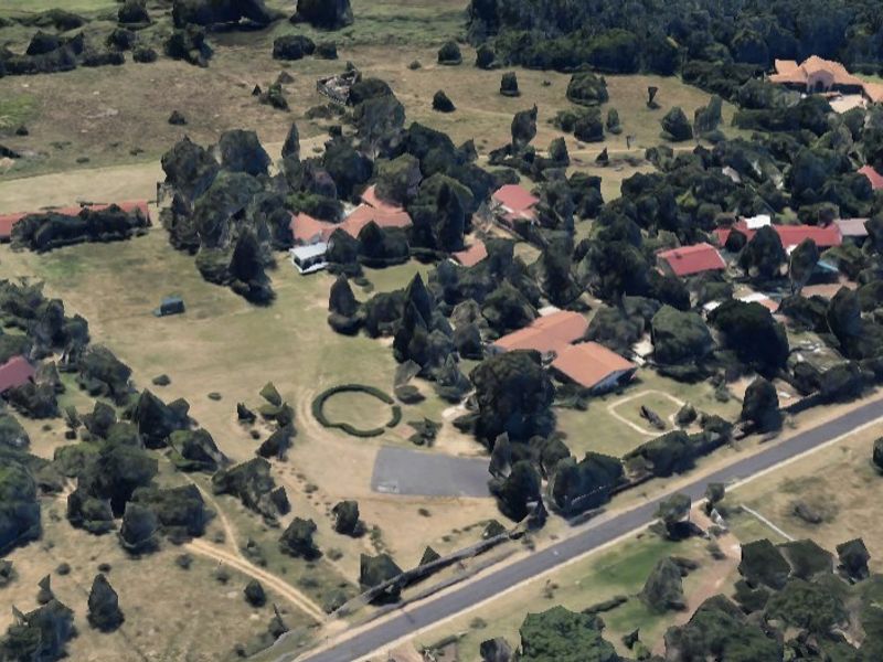 An aerial view of a residential area featuring multiple buildings, mature trees, and surrounding open grassland.
