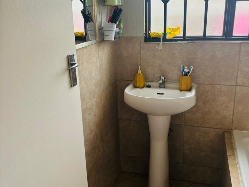 A functional bathroom featuring a pedestal basin, tiled walls and flooring, and a window fitted with a security gate.