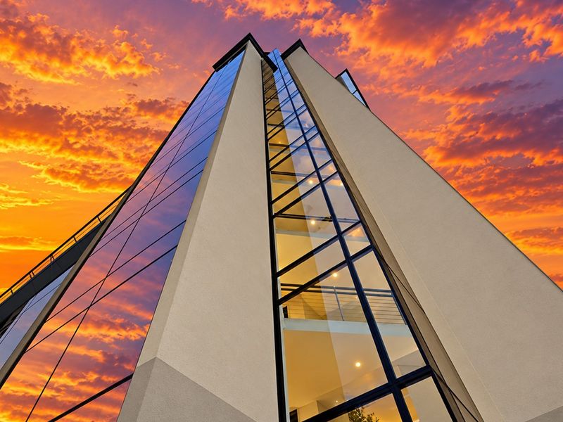 A low-angle exterior view of a modern apartment building featuring a glass curtain wall and stucco facade against a vibrant sunset sky.