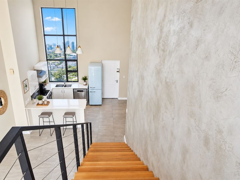 Modern open-plan kitchen and living area featuring high ceilings, large windows with city views, sleek cabinetry, and a striking textured feature wall.