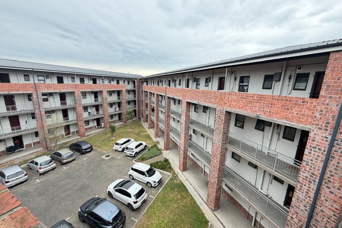 An elevated view of an apartment complex courtyard featuring exposed brick architecture, metal balcony railings, a paved parking area, and a communal garden space.