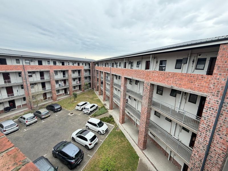 An elevated view of an apartment complex courtyard featuring exposed brick architecture, metal balcony railings, a paved parking area, and a communal garden space.