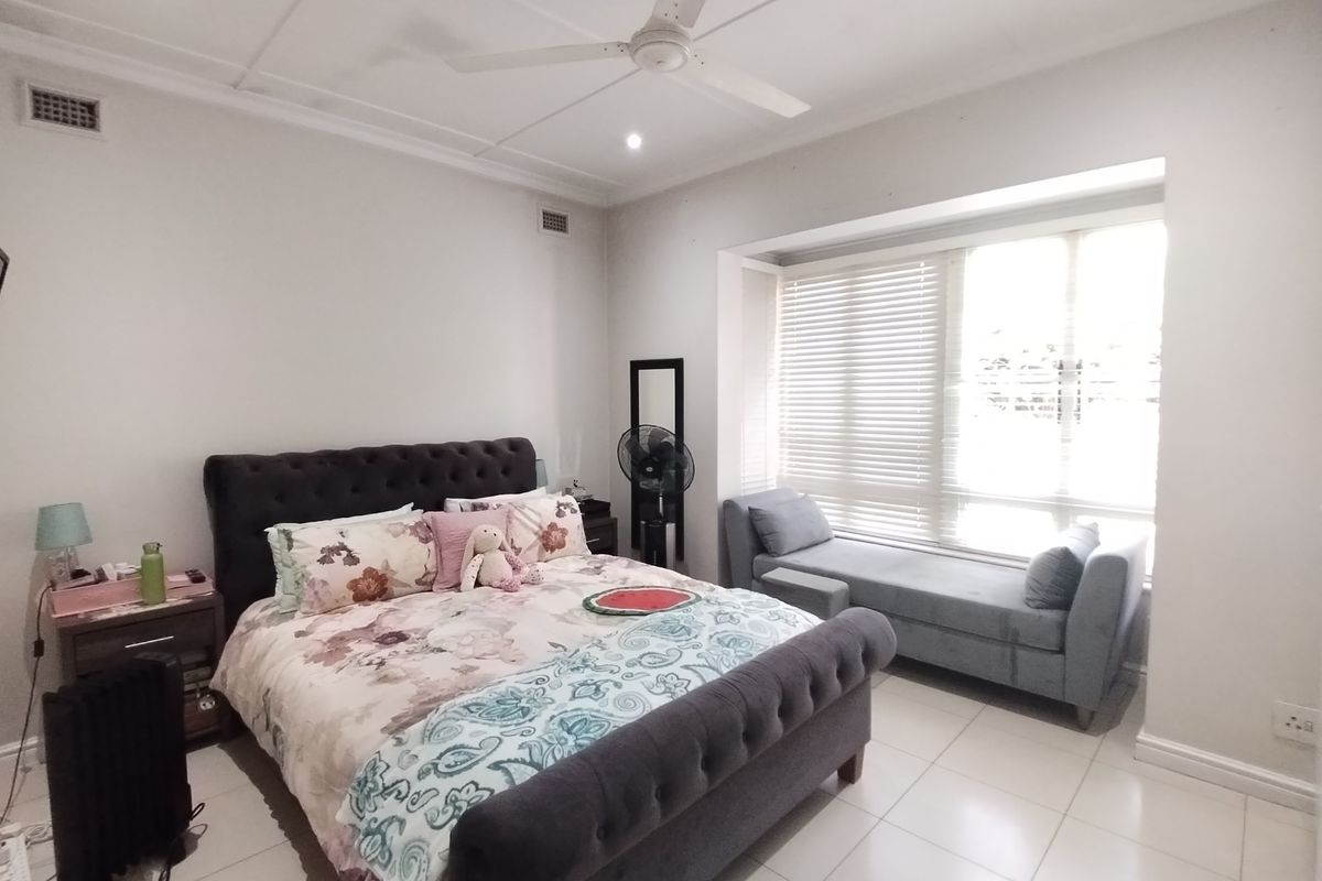 A well-lit bedroom featuring tiled flooring, a ceiling fan, and a large window with venetian blinds, providing a bright and comfortable living space.