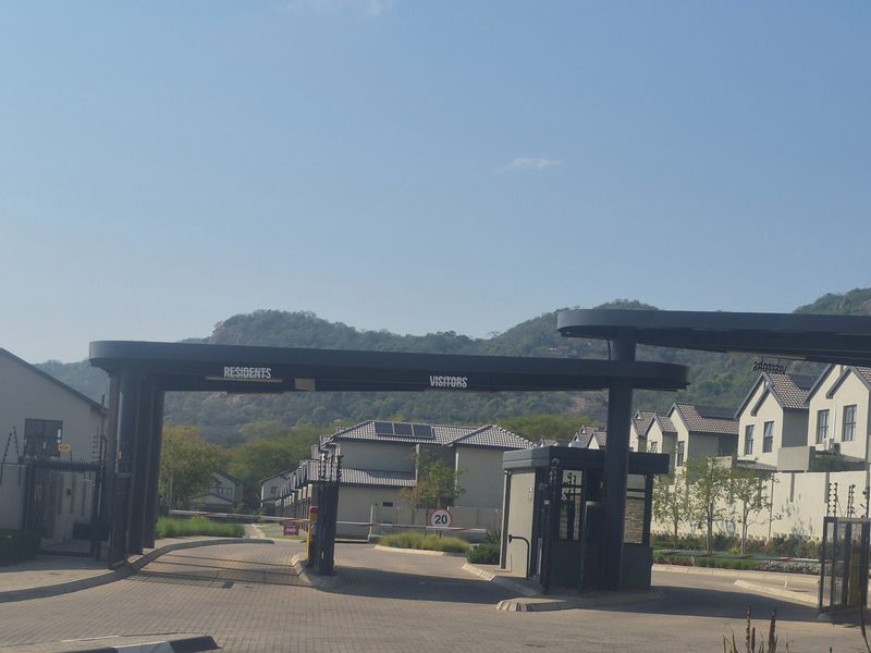 Entrance to a secure residential estate featuring a modern gatehouse, boom gates for residents and visitors, and paved access roads.