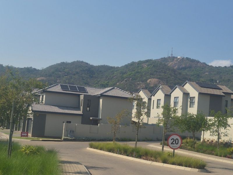 A street view of a modern residential estate featuring multi-story homes with tiled roofs, solar panels, and landscaped surroundings against a mountain backdrop.