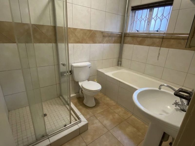 A functional bathroom featuring a glass shower enclosure, a built-in bathtub, a pedestal basin, and a toilet with neutral wall and floor tiling.