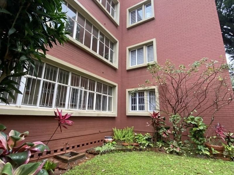 Rear view of a multi-story apartment building featuring red brick facade, large casement windows with security bars, and a well-maintained garden area.