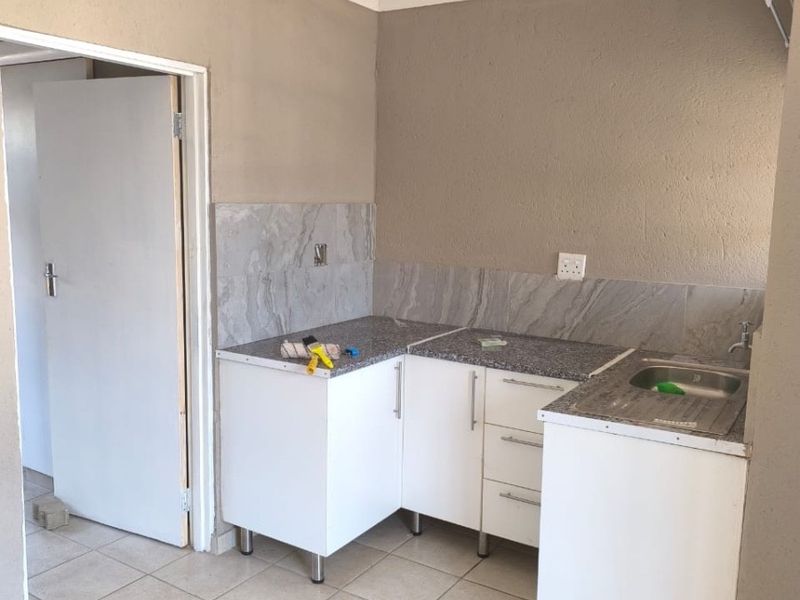 A compact, empty kitchen area featuring white base cabinetry, stone countertops, a stainless steel sink, and neutral tiled flooring with a tiled backsplash.