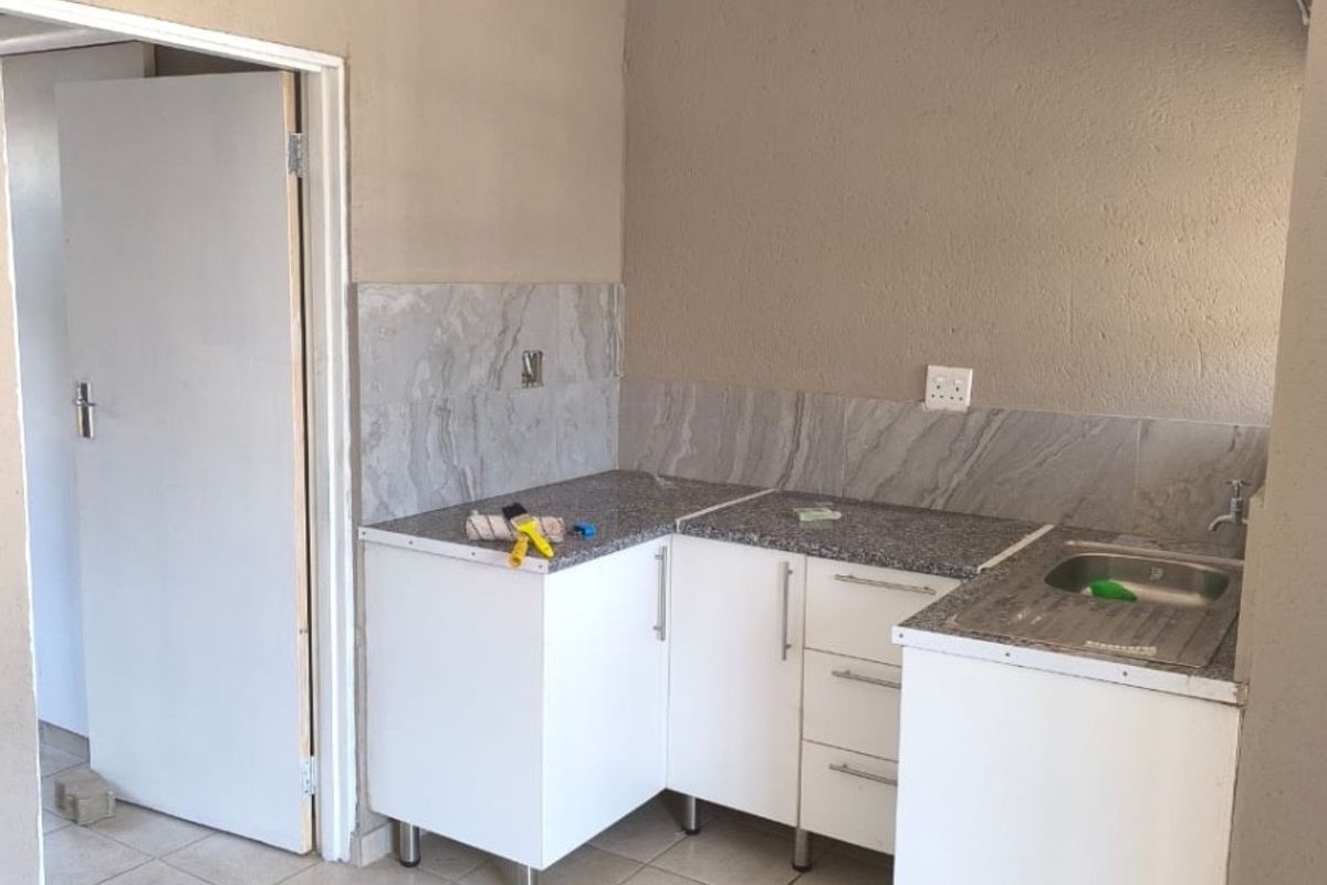A compact, empty kitchen area featuring white base cabinetry, stone countertops, a stainless steel sink, and neutral tiled flooring with a tiled backsplash.