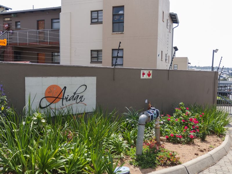 Front entrance view of the St Aidan apartment complex featuring a landscaped garden, perimeter wall with electric fencing, and paved driveway access.